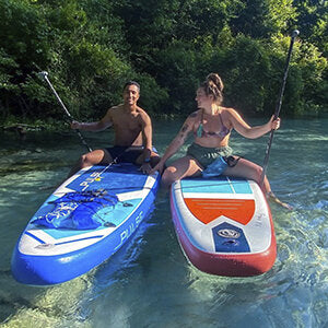 A man is riding on Pulse The Tropic 13 ft Inflatable Stand Up Paddleboard with a women ridnng Pulse The Tropic 10.6ft Inflatable Stand Up Paddleboard